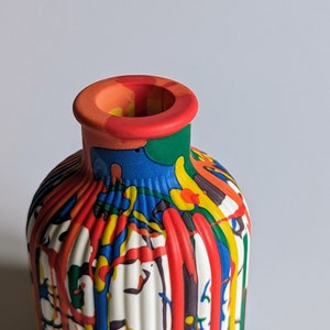 May include: A decorative ceramic vase with a white, ribbed body and a colourful, dripping paint design. The paint includes red, yellow, blue, green, and orange. The rim is red and orange. The vase is suitable for home decor.