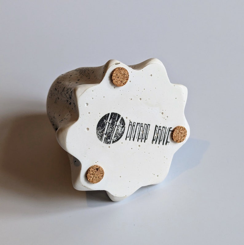 May include: White ceramic planter with a speckled finish and a starburst shape. The planter has a cork bottom and the words "Bamboo Grove" printed on the bottom.