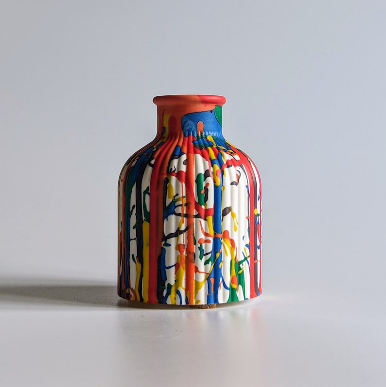 May include: A white ceramic vase with vertical ridges and a colourful paint drip design. The paint drips in red, blue, yellow, and green, creating a vibrant and artistic look. The vase has a rounded body and a narrow neck.
