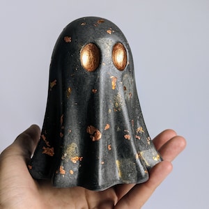 May include: A dark grey ghost ornament with gold leaf detailing. The ghost features large, circular, gold-coloured eyes. The object is held in a hand against a plain background. A unique Halloween or spooky season decoration.