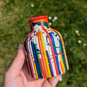 May include: A small, white ceramic vase with vertical ridges and a vibrant paint splatter design. The vase has a red rim and is decorated with streaks of red, orange, yellow, green, blue, and purple paint. The vase is held against a backdrop of green grass.