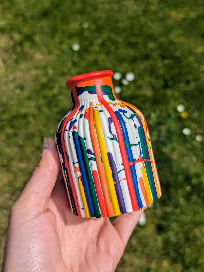 May include: A small, white ceramic vase with vertical ridges and a vibrant paint splatter design. The vase has a red rim and is decorated with streaks of red, orange, yellow, green, blue, and purple paint. The vase is held against a backdrop of green grass.