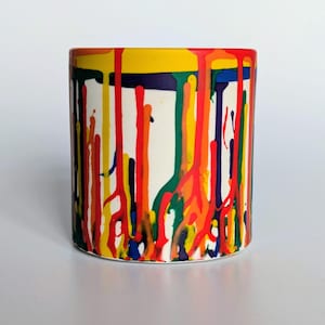 May include: A cylindrical ceramic mug with a white base, decorated with vertical streaks of bright colours including red, yellow, green, and blue. The rim is yellow, with a dark blue band beneath it. The design gives the impression of paint dripping down the sides.