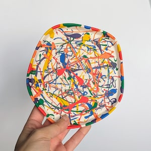 May include: A white ceramic plate with a colourful abstract design. The plate has a green, blue, red, yellow, and orange paint splatter design.