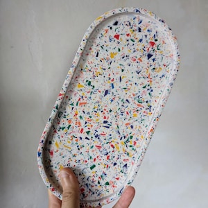 May include: An oval-shaped tray featuring a white base with a multi-coloured terrazzo pattern. The surface is speckled with small, irregular fragments in shades of red, blue, yellow, and green, creating a vibrant, confetti-like appearance.
