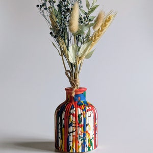 May include: A vibrant, hand-painted ceramic vase with a striped design in red, blue, yellow, and white. The vase contains a bouquet of dried flowers, including wheat, bunny tails, and small blue-green flowers, tied with twine.