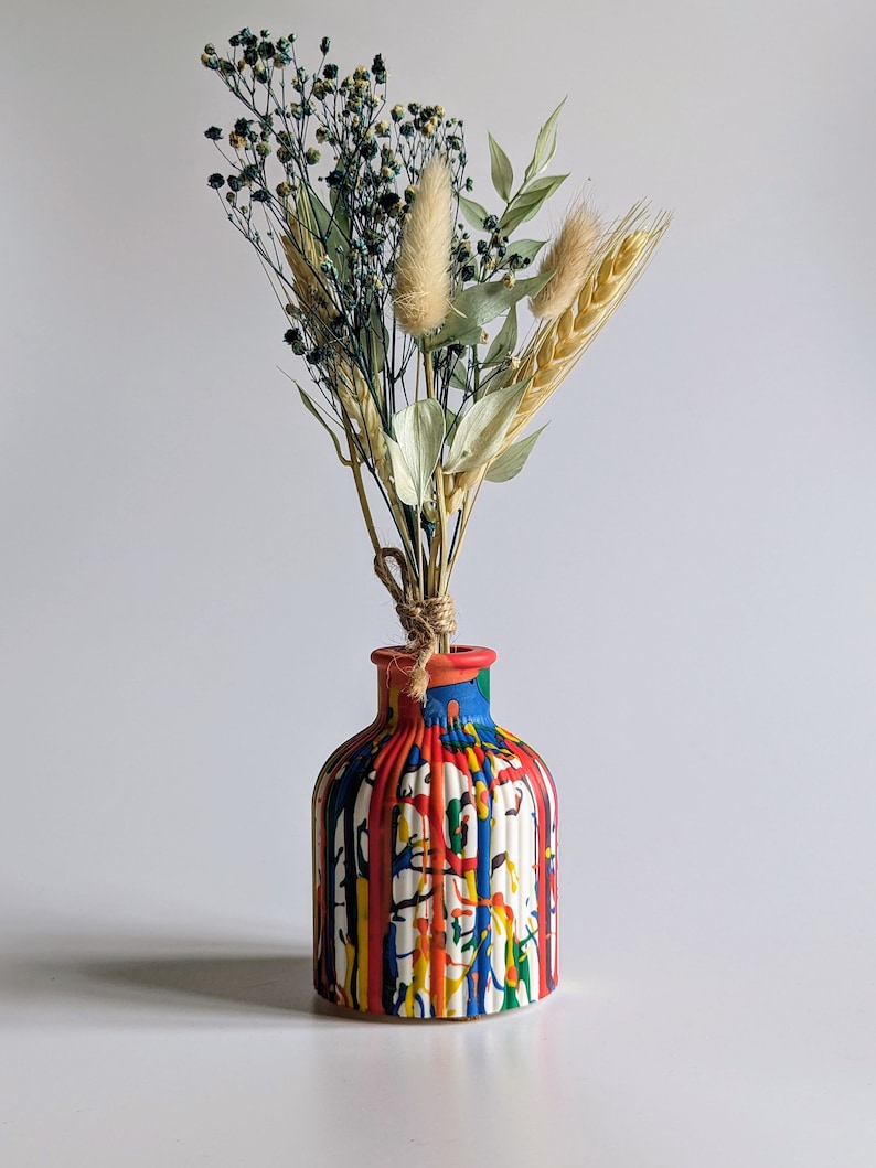 May include: A vibrant, hand-painted ceramic vase with a striped design in red, blue, yellow, and white. The vase contains a bouquet of dried flowers, including wheat, bunny tails, and small blue-green flowers, tied with twine.
