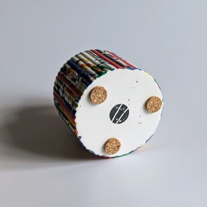 May include: A cylindrical planter with a textured, colourful exterior and a white base. The base has three cork feet and a black circular stamp. The planter's exterior features vertical stripes of various colours.