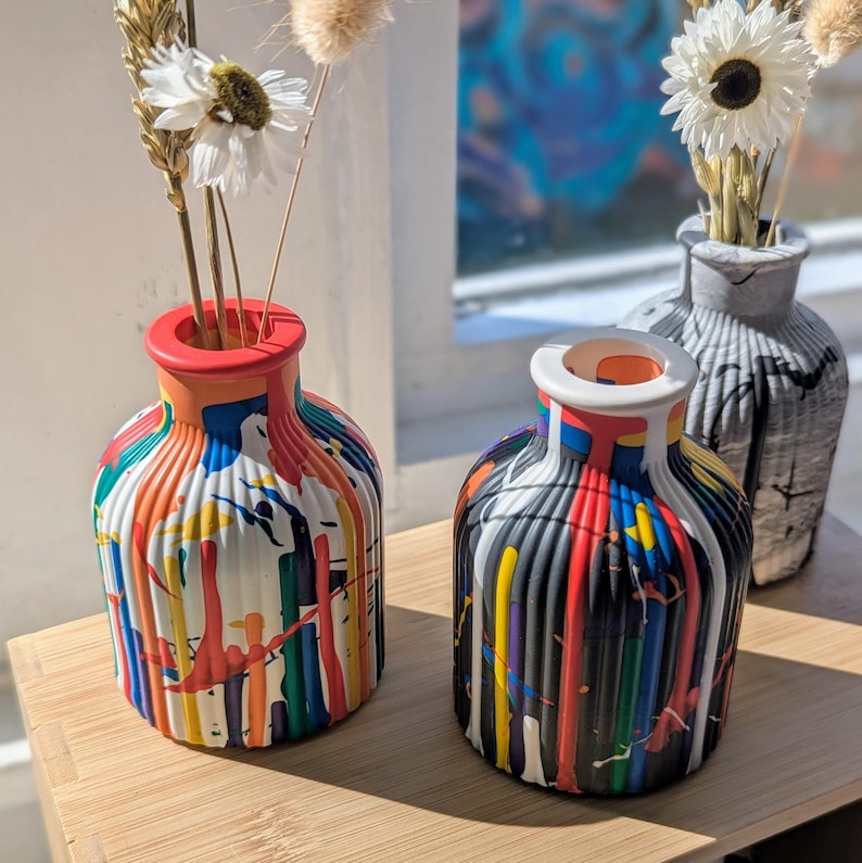 May include: Three decorative vases with dried flowers. The two front vases have a colourful paint drip design in red, yellow, blue, and black. The third vase is grey with vertical ridges. The vases are on a wooden surface.
