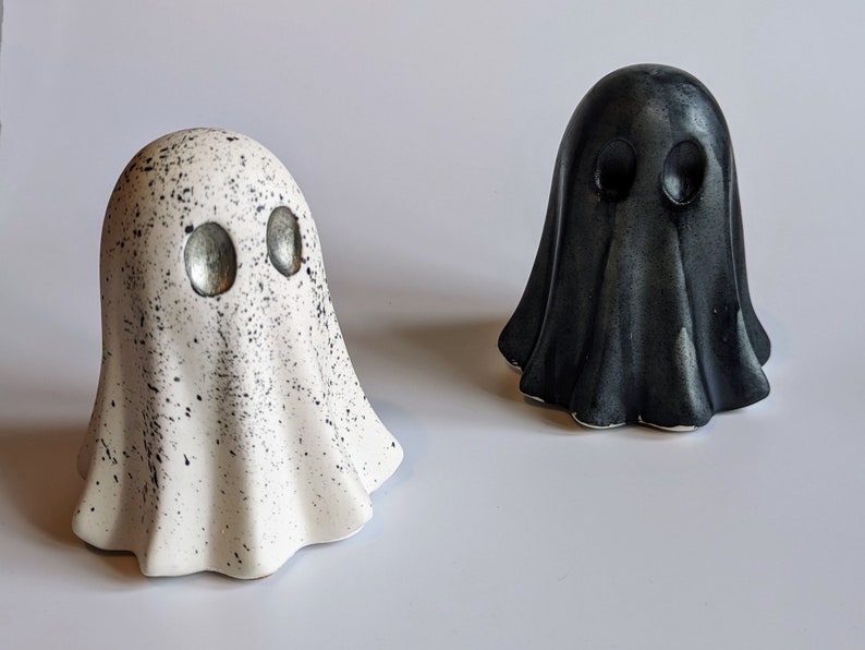 May include: Two ceramic ghost figurines, one white with black speckles and silver eyes, the other black with silver eyes.