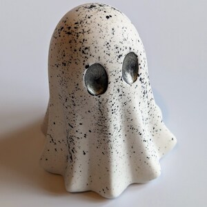 May include: A white ceramic ghost figurine with black splattered paint and silver eyes. The ghost has a rounded head and a flowing, ruffled body.