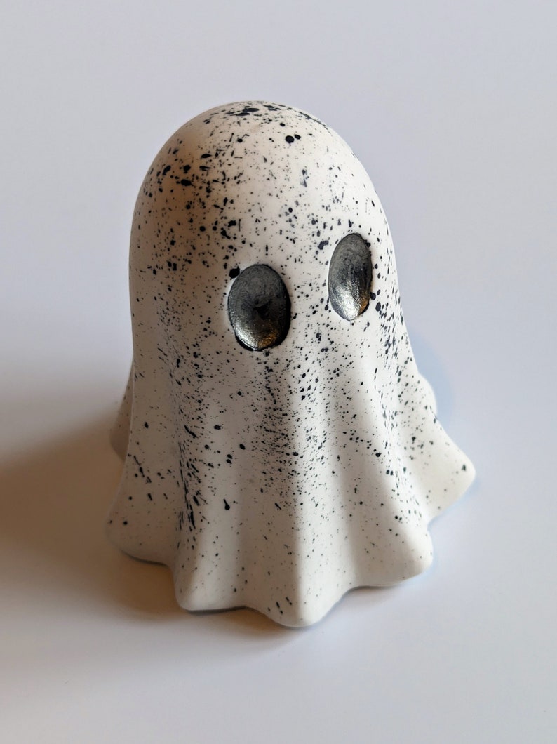 May include: A white ceramic ghost figurine with black splattered paint and silver eyes. The ghost has a rounded head and a flowing, ruffled body.