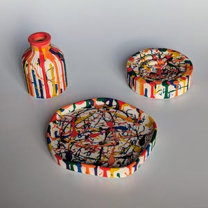 May include: A trio of ceramic items with a white base and a vibrant paint splatter design. The set comprises a small vase, a circular dish, and an oval tray. The vase features a red rim, and the paintwork incorporates red, blue, yellow, and green.
