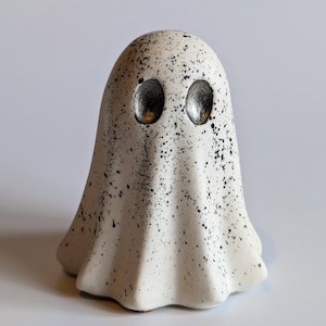 May include: A white ceramic ghost figurine with black speckles and silver eyes. The ghost has a rounded head and a flowing, draped body.
