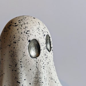 May include: A white ceramic ghost figurine with black speckles and silver eyes. The ghost has a rounded head and a flowing, draped body.