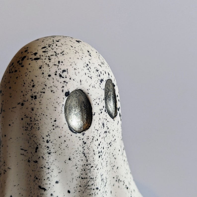 May include: A white ceramic ghost figurine with black speckles and silver eyes. The ghost has a rounded head and a flowing, draped body.