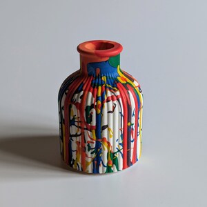 May include: A small, white ceramic vase with a colourful paint drip design. The vase has a round body with vertical ridges and a narrow neck. The paint drips are red, yellow, blue, and green.
