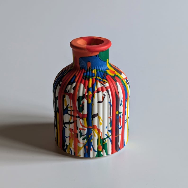 May include: A small, white ceramic vase with a colourful paint drip design. The vase has a round body with vertical ridges and a narrow neck. The paint drips are red, yellow, blue, and green.