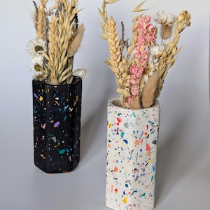 May include: Two decorative vases with dried flowers. One vase is black with colorful flecks, the other is white with colorful flecks. The vases are rectangular with a geometric shape and contain dried wheat, daisies, and other dried flowers.
