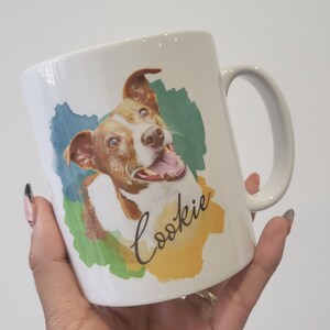 May include: White ceramic mug featuring a watercolour design of a brown and white dog with blue eyes. The dog is smiling and the text "Cookie" is written in cursive on the mug.