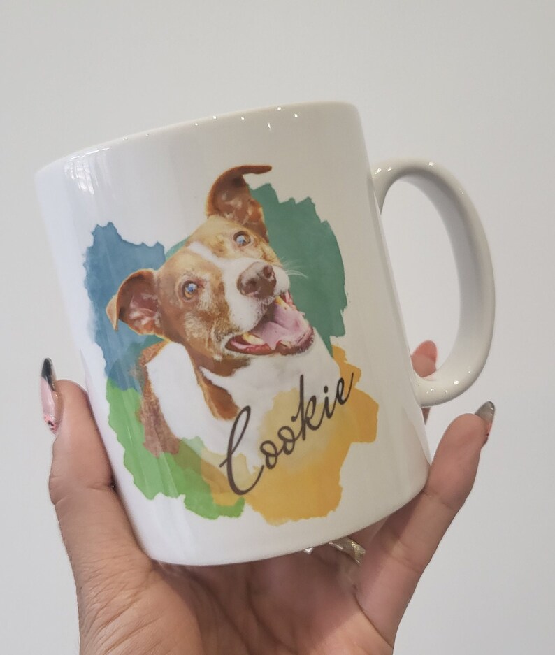 May include: White ceramic mug featuring a watercolour design of a brown and white dog with blue eyes. The dog is smiling and the text "Cookie" is written in cursive on the mug.