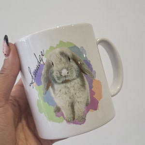 May include: White ceramic mug featuring a watercolour design of a grey bunny with floppy ears. The bunny is surrounded by colourful watercolour splatters. The mug has the word "Snudge" written on the side in black cursive.