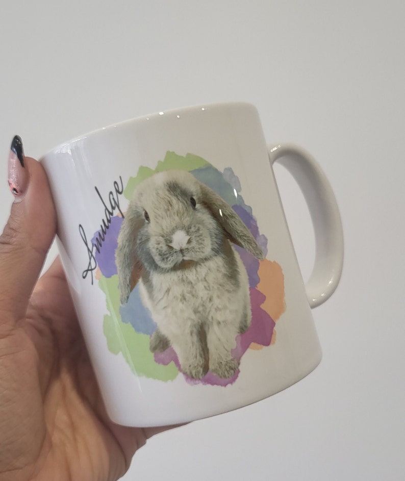 May include: White ceramic mug featuring a watercolour design of a grey bunny with floppy ears. The bunny is surrounded by colourful watercolour splatters. The mug has the word "Snudge" written on the side in black cursive.