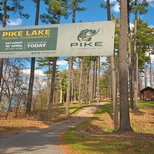 May include: A white sign with green and yellow text announcing a Pike Lake Fishing Tournament on Saturday, 10th April from 10 am to 4 pm. The sign is hanging between two trees in a wooded area with a dirt path leading into the woods.