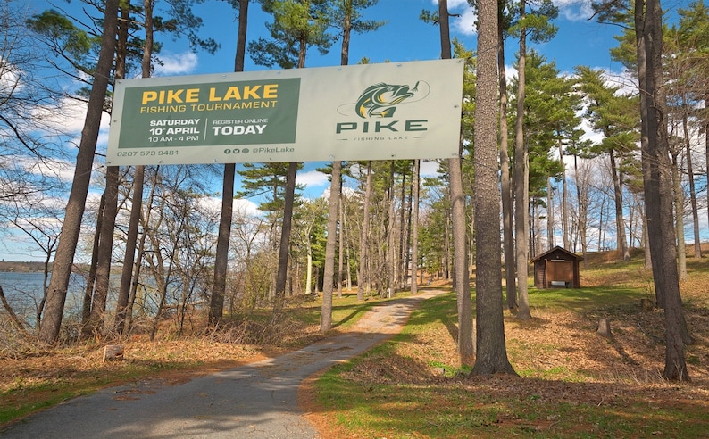 May include: A white sign with green and yellow text announcing a Pike Lake Fishing Tournament on Saturday, 10th April from 10 am to 4 pm. The sign is hanging between two trees in a wooded area with a dirt path leading into the woods.