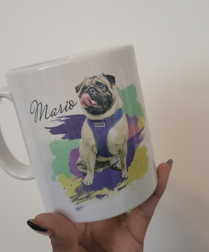 May include: White ceramic mug with a colourful abstract design featuring a pug dog wearing a purple harness. The dog's name, "Mario", is written in black cursive on the mug.