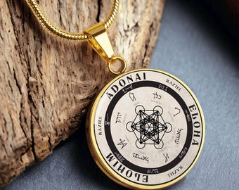 May include: Gold-toned pendant necklace with a circular charm. The charm features black text and geometric designs on a cream background, framed by a gold border. The necklace is displayed on a wooden surface.