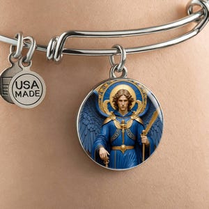 May include: Silver-tone bangle bracelet with a round charm featuring an illustration of an angel in blue robes and wings, holding a sword. The charm has a gold halo and accents. A smaller charm reads "USA MADE".