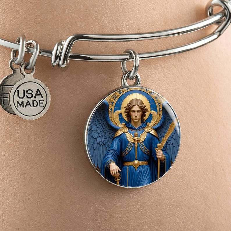 May include: Silver-tone bangle bracelet with a round charm featuring an illustration of an angel in blue robes and wings, holding a sword. The charm has a gold halo and accents. A smaller charm reads "USA MADE".