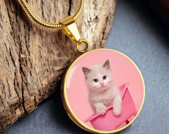 May include: Gold-colored pendant necklace featuring a round charm with a white kitten in a pink bathtub against a pink background. The kitten has blue eyes and gray ears. The necklace is displayed on a wooden surface.
