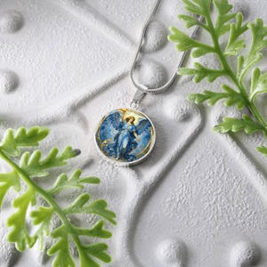 K&ouml;nnte beinhalten: Eine silberfarbene Halskette mit einem runden Anh&auml;nger, der einen blauen Engel mit ausgebreiteten Fl&uuml;geln vor einem goldenen und blauen Hintergrund zeigt. Die Halskette wird auf einer strukturierten wei&szlig;en Oberfl&auml;che mit gr&uuml;nem Laub im Vordergrund pr&auml;sentiert.