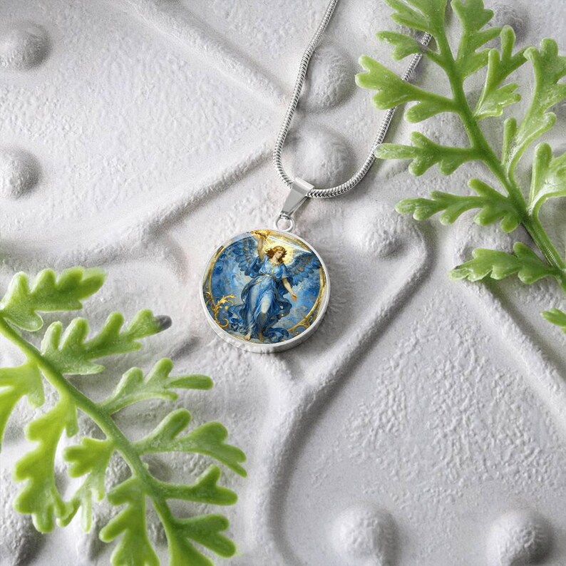 K&ouml;nnte beinhalten: Eine silberfarbene Halskette mit einem runden Anh&auml;nger, der einen blauen Engel mit ausgebreiteten Fl&uuml;geln vor einem goldenen und blauen Hintergrund zeigt. Die Halskette wird auf einer strukturierten wei&szlig;en Oberfl&auml;che mit gr&uuml;nem Laub im Vordergrund pr&auml;sentiert.