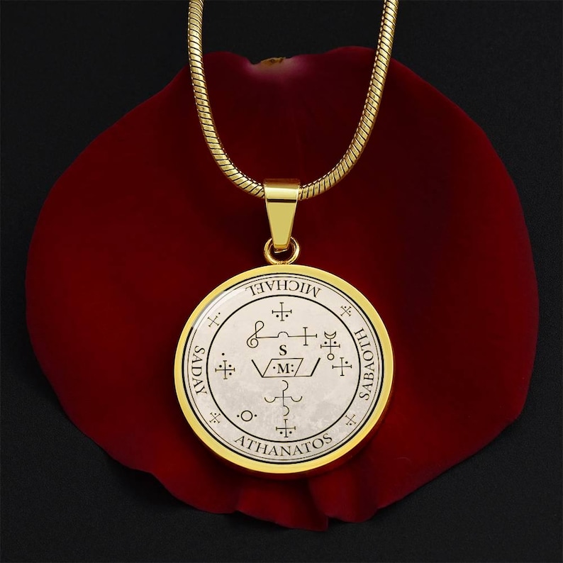 May include: A gold-colored pendant necklace with a circular charm featuring intricate symbols and the name "MICHAEL". The pendant is set against a dark red background, with a gold chain. The charm also includes the words "SADAY", "SABATH", and "ATHANATOS".
