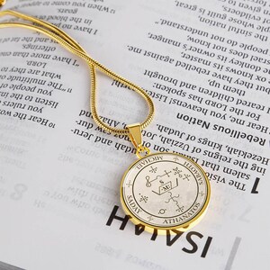 May include: A gold-colored pendant necklace with a circular charm. The charm features an intricate design with symbols and text, including the name "MICHAEL". The necklace rests on a book page with printed text.