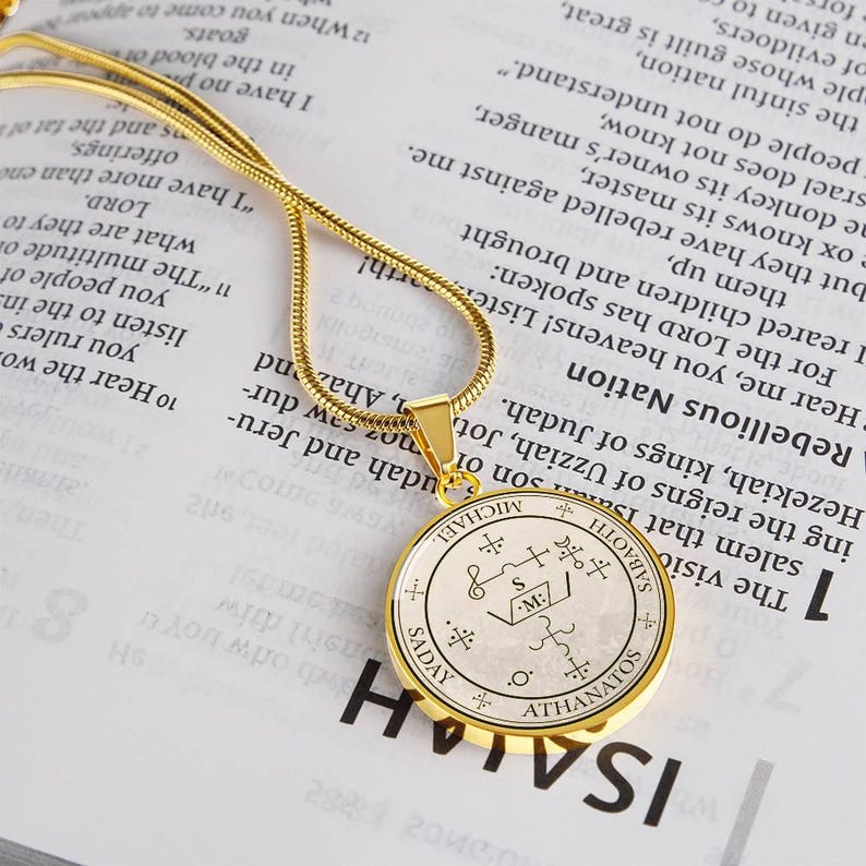 May include: A gold-colored pendant necklace with a circular charm. The charm features an intricate design with symbols and text, including the name "MICHAEL". The necklace rests on a book page with printed text.