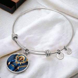 May include: A silver-colored bangle bracelet with a round charm featuring an angel in blue robes. The charm has a gold halo and is attached to the bracelet with a small ring. Two smaller charms are also attached, one with the text "HAND MADE" and another with a flag design.