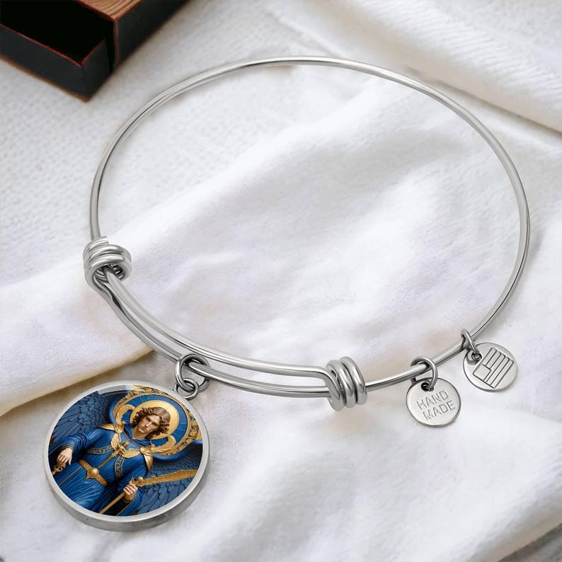 May include: A silver-colored bangle bracelet with a round charm featuring an angel in blue robes. The charm has a gold halo and is attached to the bracelet with a small ring. Two smaller charms are also attached, one with the text "HAND MADE" and another with a flag design.