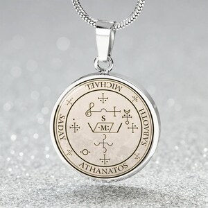 May include: Silver-toned pendant necklace with a circular charm. The charm features intricate symbols and text, including the words "MICHAEL", "SADAY", "SABAOTH", and "ATHANATOS". The pendant hangs from a delicate silver chain.