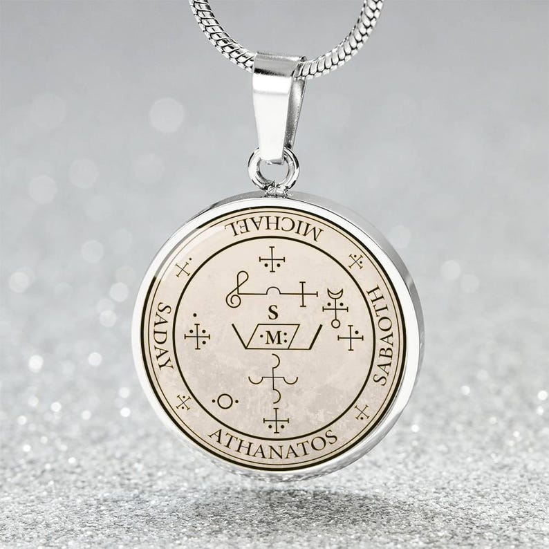 May include: Silver-toned pendant necklace with a circular charm. The charm features intricate symbols and text, including the words "MICHAEL", "SADAY", "SABAOTH", and "ATHANATOS". The pendant hangs from a delicate silver chain.