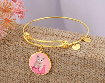 May include: Gold-colored bangle bracelet with a round charm featuring a kitten in a pink tub. The charm is suspended from the bracelet, which has a simple, elegant design. Additional small gold charms are attached to the bracelet.