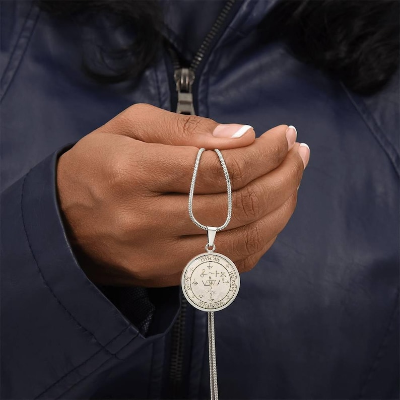 May include: A silver-colored pendant necklace with a circular charm. The charm features intricate symbols and text. The necklace is held against a dark blue jacket. The pendant hangs on a thin, silver chain.