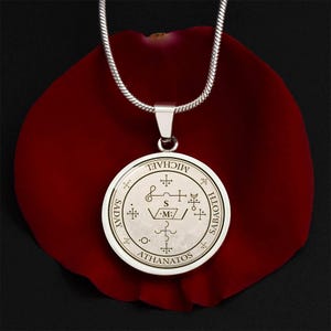 May include: Silver-tone pendant necklace with a round charm featuring intricate symbols and the words "MICHAEL", "SADAY", "SABATH", and "ATHANATOS". The pendant hangs on a silver chain, set against a red background.