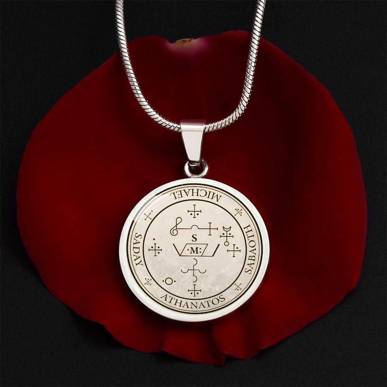 May include: Silver-tone pendant necklace with a round charm featuring intricate symbols and the words "MICHAEL", "SADAY", "SABATH", and "ATHANATOS". The pendant hangs on a silver chain, set against a red background.