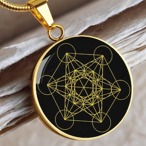 May include: Gold-tone pendant necklace featuring a black circular pendant with a yellow geometric design. The pendant hangs from a gold chain. The design includes circles, lines, and geometric shapes. The necklace is displayed on a textured surface.