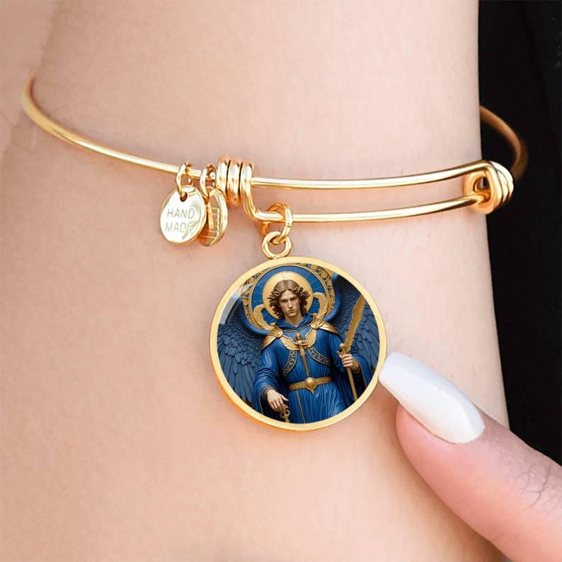 May include: Gold-colored bangle bracelet with a round pendant featuring an illustration of an angel in blue robes, holding a sword. The pendant is framed in gold and has a blue halo. Small gold charms are attached to the bracelet, one reading "HAND MADE".