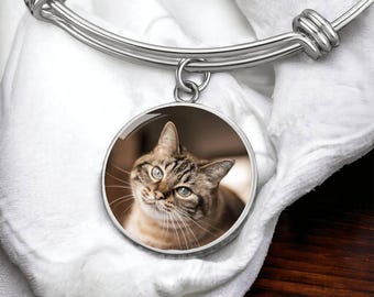 May include: A silver-toned bangle bracelet with a round pendant featuring a cat. The cat has brown and tan fur with blue eyes. The bracelet is resting on a white, textured surface.